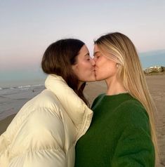 two women are kissing on the beach while one woman is wearing a green sweater and white jacket