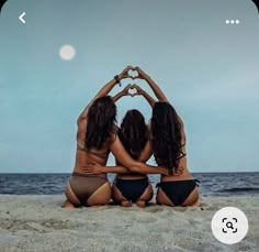 three women in bikinis sitting on the beach making a heart shape with their hands