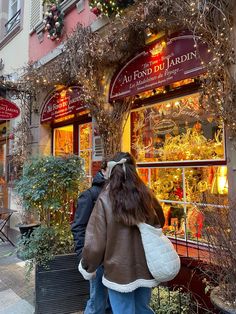 two people walking past a store front with christmas decorations on the windows and trees around it