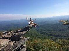 two people sitting on the edge of a cliff with their arms in the air above them