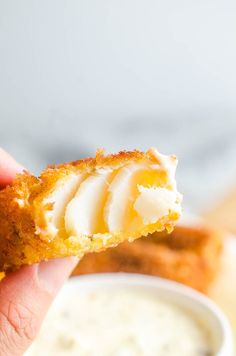 a person holding up a piece of food with cream on it and dipping sauce in the background