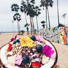 a person holding up a coconut bowl filled with fruit and nuts on top of a sandy beach