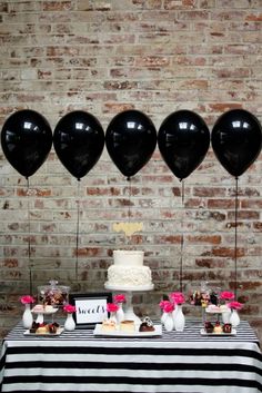 a table topped with black balloons next to a cake and cupcakes in front of a brick wall