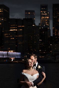 a bride and groom pose for a photo in front of the city skyline at night