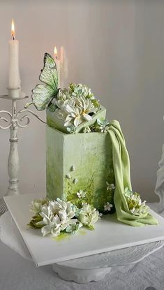 a green cake with white flowers and butterflies on it, sitting on a table next to two candles