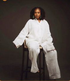 a woman sitting on top of a wooden chair wearing white pants and a long sleeved shirt