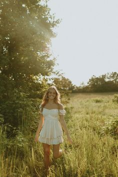 a woman in a white dress is walking through tall grass and trees with her hands behind her back