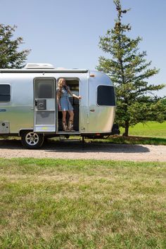 a woman is standing in the open door of a silver trailer parked on a gravel road