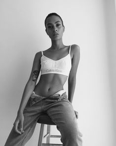 a woman sitting on top of a wooden stool next to a white wall and wearing jeans