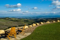 two wooden chairs sitting next to each other on top of a grass covered hillside with rolling hills in the background