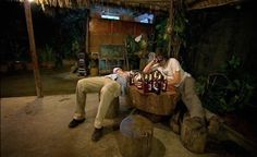 two men are sitting at a table with bottles on it and one man is drinking from a bottle