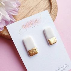 white and gold square stud earrings sitting on top of a pink table next to a flower