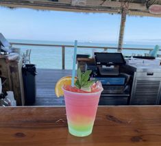 a pink and green drink sitting on top of a wooden table next to the ocean