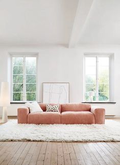 a living room with white walls and wooden floors, an orange couch is in front of two windows