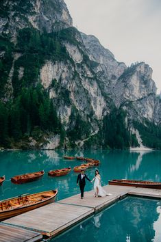 a bride and groom walking on a dock next to boats in the water with mountains in the background