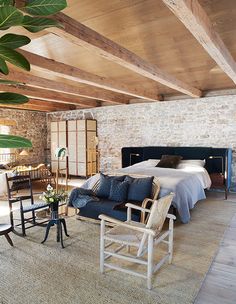 a bedroom with stone walls and exposed ceiling, blue bedding, two armchairs, an end table in the foreground