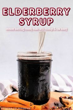 A photo of a glass mason jar full of elderberry syrup with a silver spoon inside the jar, sitting on top of a wooden cutting board with cinnamon sticks and a tablespoon full of dried rose hips on it, all sitting next to a striped kitchen towel with text that says Elderberry Syrup Immune Boosting Remedy Made with Rosehips & Raw Honey.