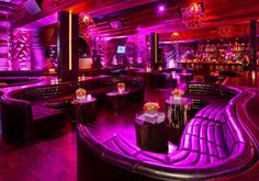 a dimly lit room with purple lighting and black leather couches, tables and stools