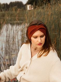 a woman wearing a knit headband sitting in front of a pond holding a cell phone