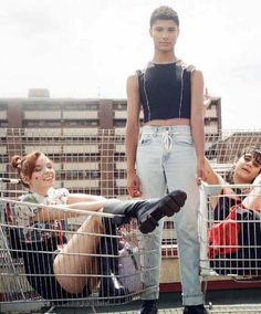 a woman standing in front of some shopping carts with her legs crossed and two other women sitting on the ground behind her