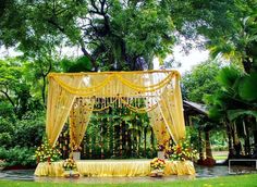 an outdoor wedding setup with yellow drapes and flowers on the bedding, surrounded by greenery