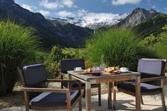 two chairs and a table with food on it in front of mountains, grass and flowers