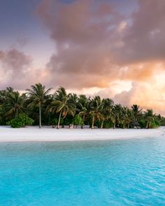 an island with palm trees and white sand in the ocean under a cloudy sky at sunset