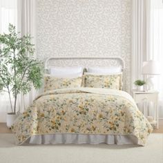 a white bed with yellow flowers on it in a room next to a window and a potted plant