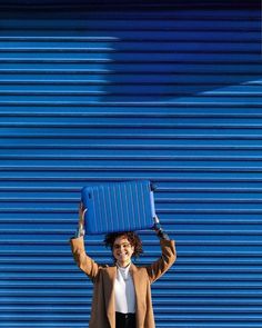 a woman holding a blue suitcase on her head while standing in front of a blue wall