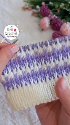 someone holding up a purple and white striped knitted object with flowers in the background