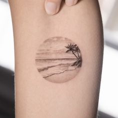 a woman's arm with a small palm tree on the beach in front of it