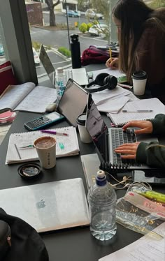 a woman sitting at a table with laptops and notebooks on it, in front of a window