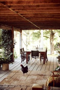 a chicken walking across a wooden deck in front of a table with chairs and potted plants