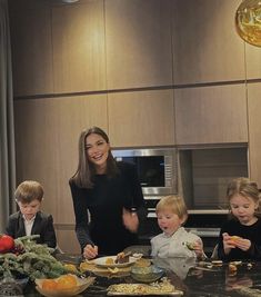 a woman standing in front of a kitchen counter with three children sitting at the table