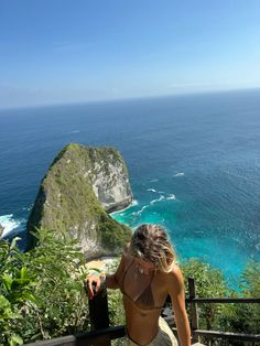 a woman sitting on top of a wooden bench looking at the ocean and cliffs in the distance