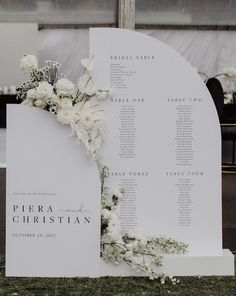 a wedding seating plan with white flowers and greenery on the back of an arch