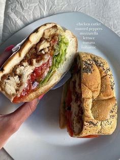 a hand holding a sandwich on top of a white plate next to a bun covered in lettuce and tomato