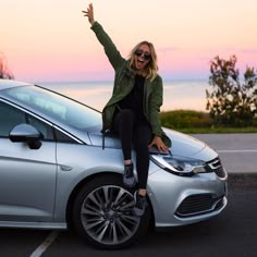 a woman sitting on the hood of a silver car with her arms in the air