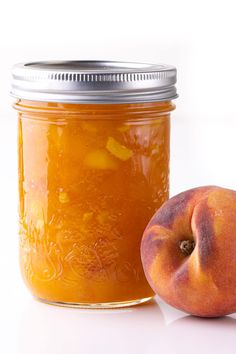 a peach jam in a glass jar next to a half - eaten peach on a white surface