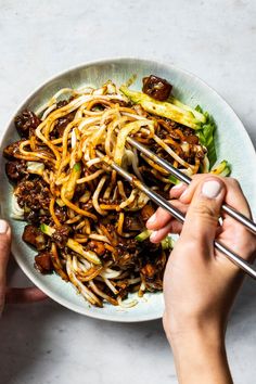 two hands holding chopsticks over a bowl of noodles and vegetables with sauce on the side