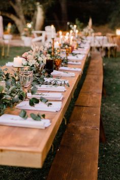 a long wooden table topped with lots of plates and glasses covered in greenery next to candles