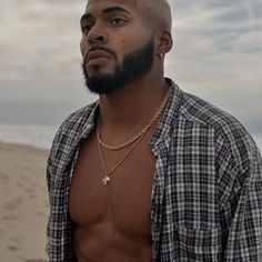 a shirtless man standing on the beach looking off into the distance with his hands in his pockets