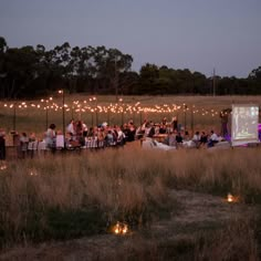 a large group of people sitting at tables in the middle of a field with lights strung over them