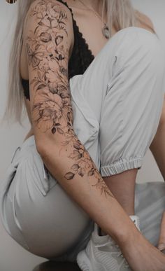 a woman sitting on the ground with her arm covered by flowers and leaves tattoo design