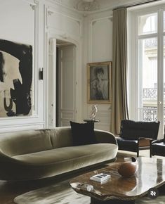 a living room with couches, chairs and paintings on the wall in front of large windows