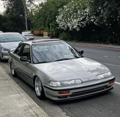 a silver car parked on the side of a road next to a sidewalk with trees in the background