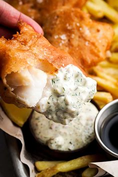 a person holding up a piece of fish and chips with dipping sauce on the side