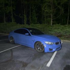 a blue car is parked in a parking lot at night with the lights on and trees behind it