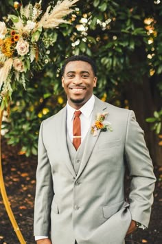 a man in a gray suit and tie standing next to a flower arch with an orange boutonniere on it