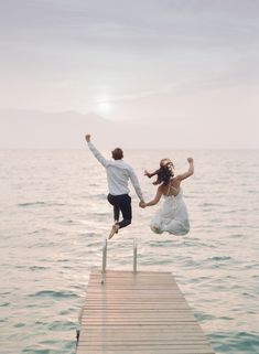 two people jumping into the air from a dock in front of some water and one person is wearing a white shirt